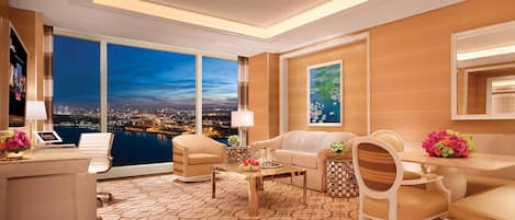A modern hotel room with a city view, a large window, a sofa, a coffee table, and a desk with a lamp.