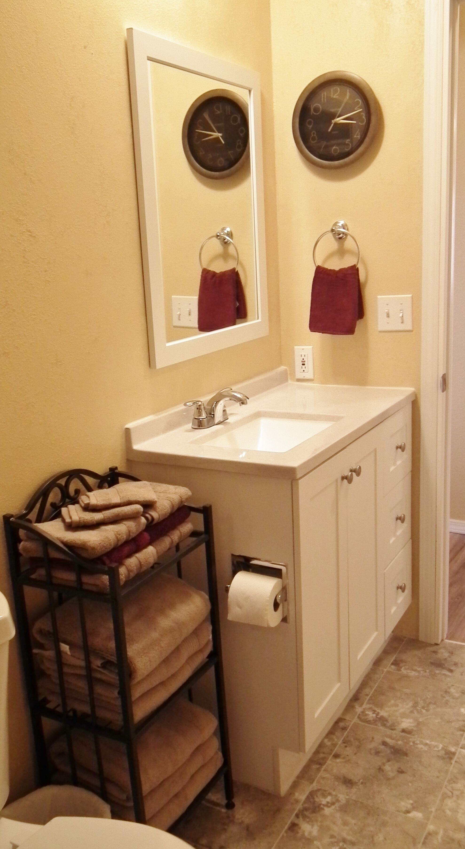 A bathroom with a white sink, a mirror, two wall clocks, a towel rack with folded towels, and a toilet paper holder.