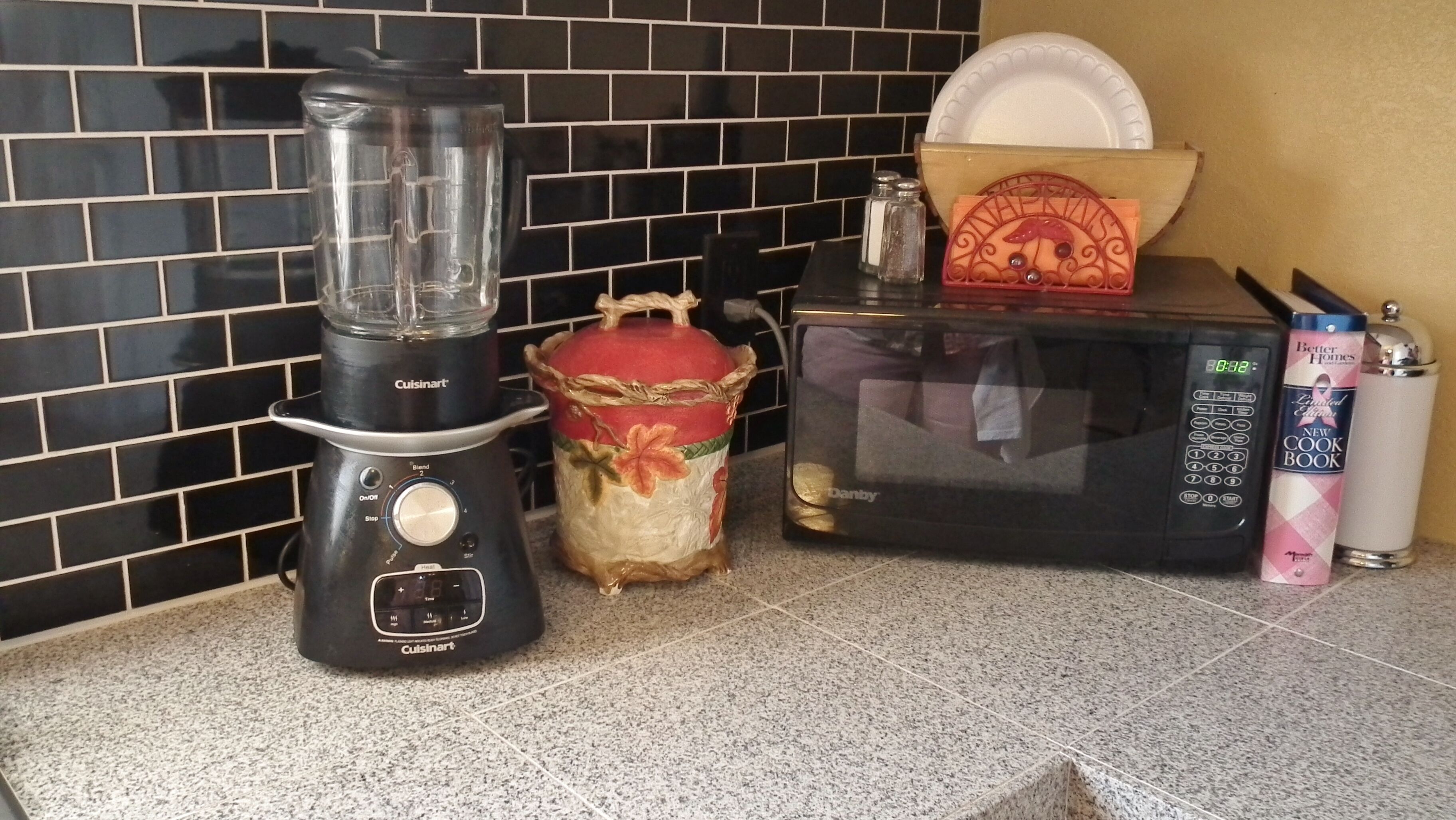 Una encimera de cocina con una licuadora Cuisinart, un microondas, un recipiente decorativo y una lata de aerosol para cocinar.