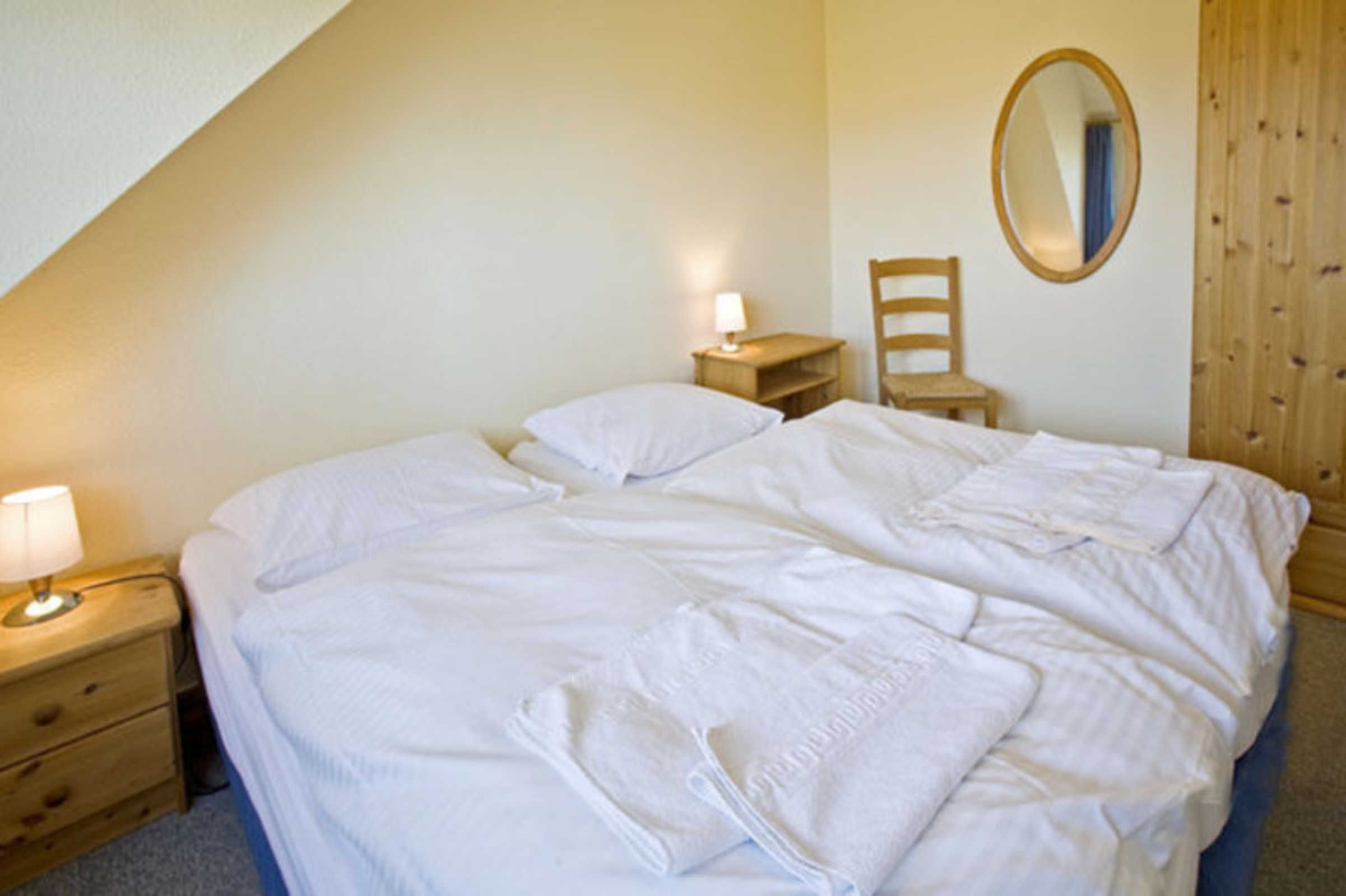 Un dormitorio con una cama, una mesita de noche con lámpara, una silla, un armario de madera y un espejo en la pared.