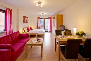 A hotel room with a bed, a red sofa, a wooden table with a bottle and glasses, a dining table with chairs, and a TV. - Apartment 2 - Apartment "am-edersee" (Edertal)