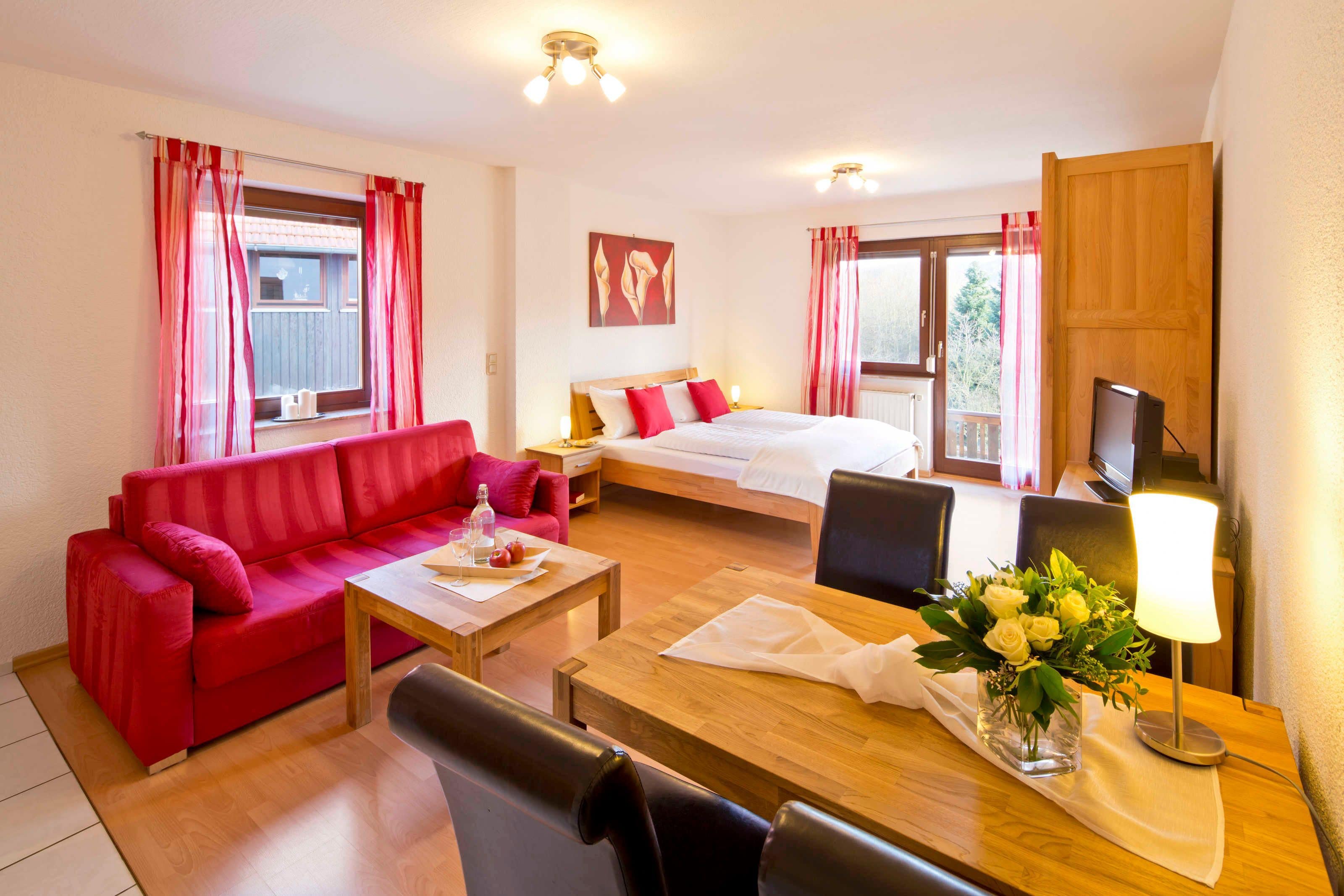 Une chambre d’hôtel avec un canapé rouge, une table à manger en bois, un lit avec une literie rouge et blanche, une télévision et une fenêtre avec des rideaux rouges.