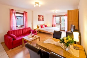A hotel room with a red sofa, a wooden dining table, a bed with red and white bedding, a TV, and a window with red curtains.