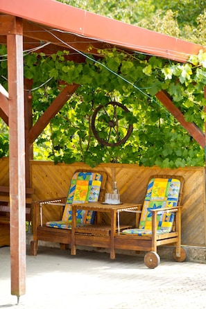 A wooden pergola with a rustic design, two wooden chairs with colorful cushions, and a metal wheel chair.