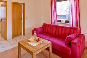 Un salon avec un canapé rouge, une table basse en bois sur laquelle se trouvent un plateau de pommes et une bouteille, et une fenêtre avec des rideaux.