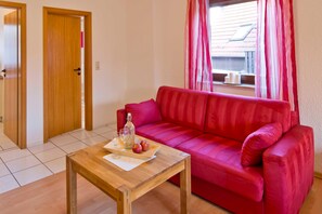 A living room with a red sofa, a wooden coffee table with a tray of apples and a bottle, and a window with curtains. - Apartment 2 - Apartment "am-edersee" (Edertal)