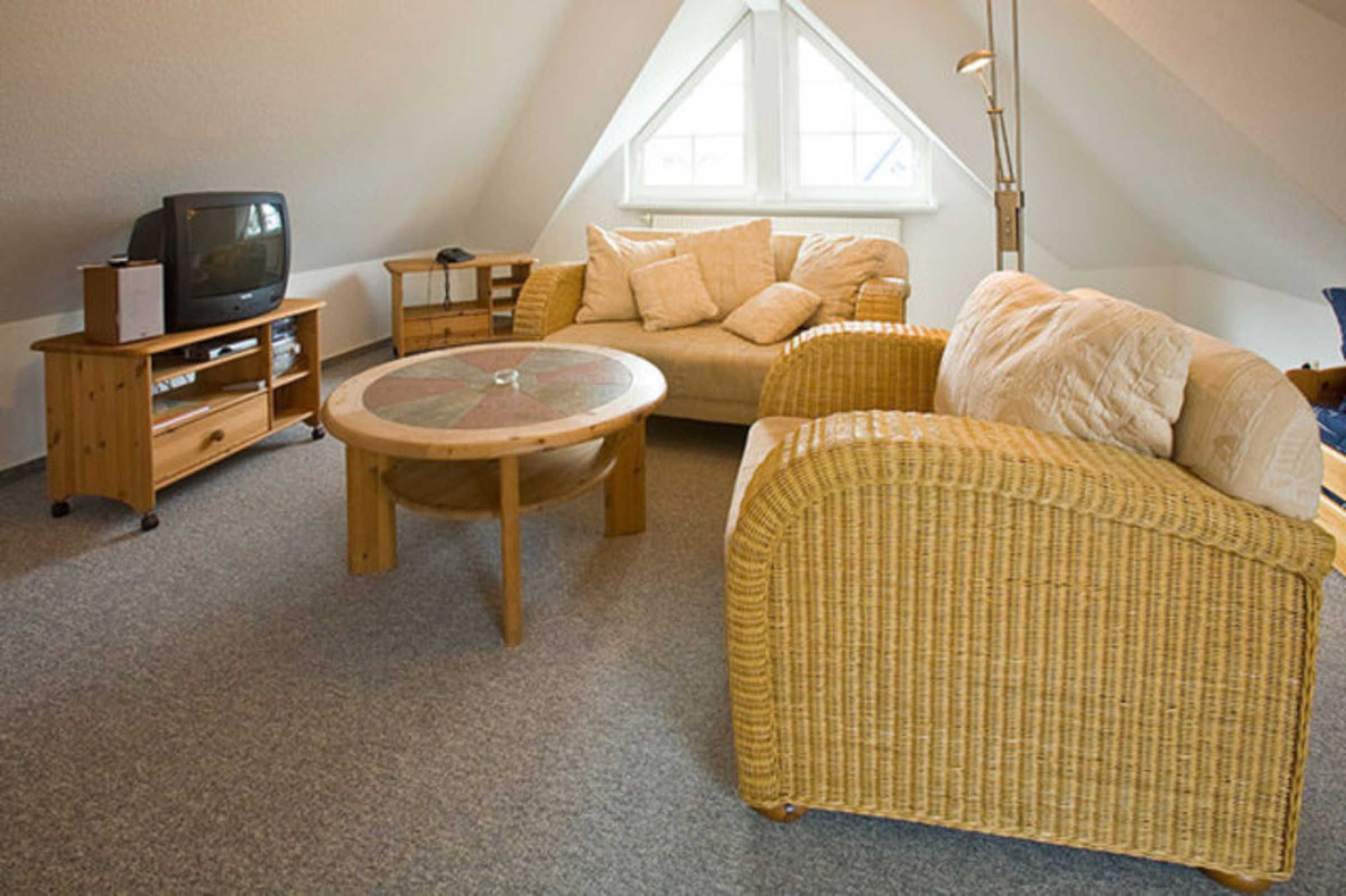 Een knus slaapkamerloft met een schuine kap, een groot raam, een rieten bank, een ronde tafel en een televisie op een houten standaard.