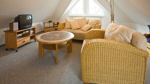 Et hyggeligt loftværelse med skråvægge, et stort vindue, en fletsofa, et rundt bord og et fjernsyn på et træstel.