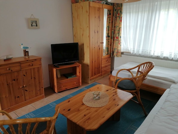 A room with a wooden wardrobe, a television on a wooden stand, a wicker chair, a small table with a glass, and a bed with white bedding.