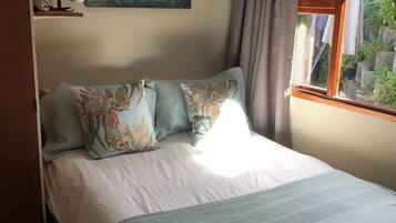 A bedroom with a bed, a painting, a window with curtains, and a shelf with a lamp.