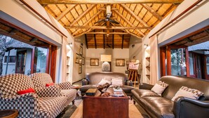A spacious living room with checkered sofas, a leather couch, and a wooden coffee table. A ceiling fan is visible, and the room has large windows on one side. - Ivory Wilderness River Rock Lodge (Bushbuckridge)