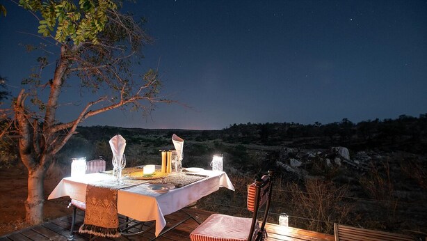 An outdoor dining setup with a table and chairs, surrounded by a natural landscape and a night sky. - Ivory Wilderness River Rock Lodge (Bushbuckridge)
