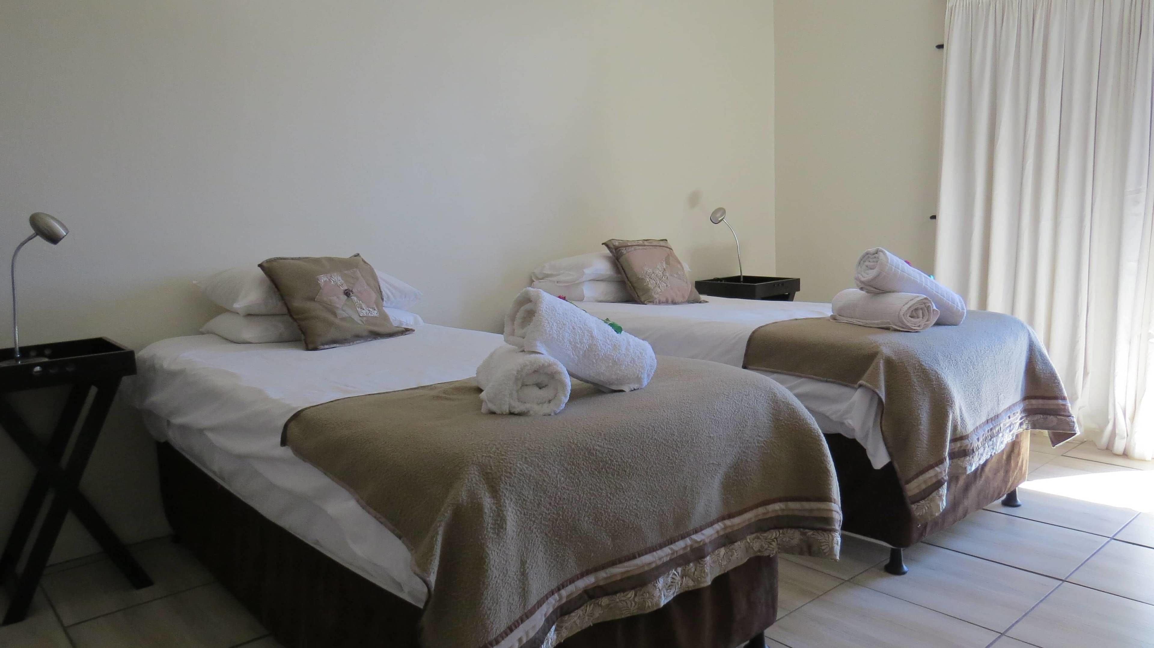 Habitación de hotel con dos camas, cada una con una toalla enrollada y una manta marrón. Se observa un mesita de noche con una lámpara y una pequeña bandeja negra.