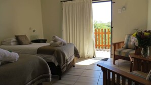 A hotel room with two beds, a wooden bench, a chair, a table, a vase with flowers, and a view of a wooden deck and greenery outside.