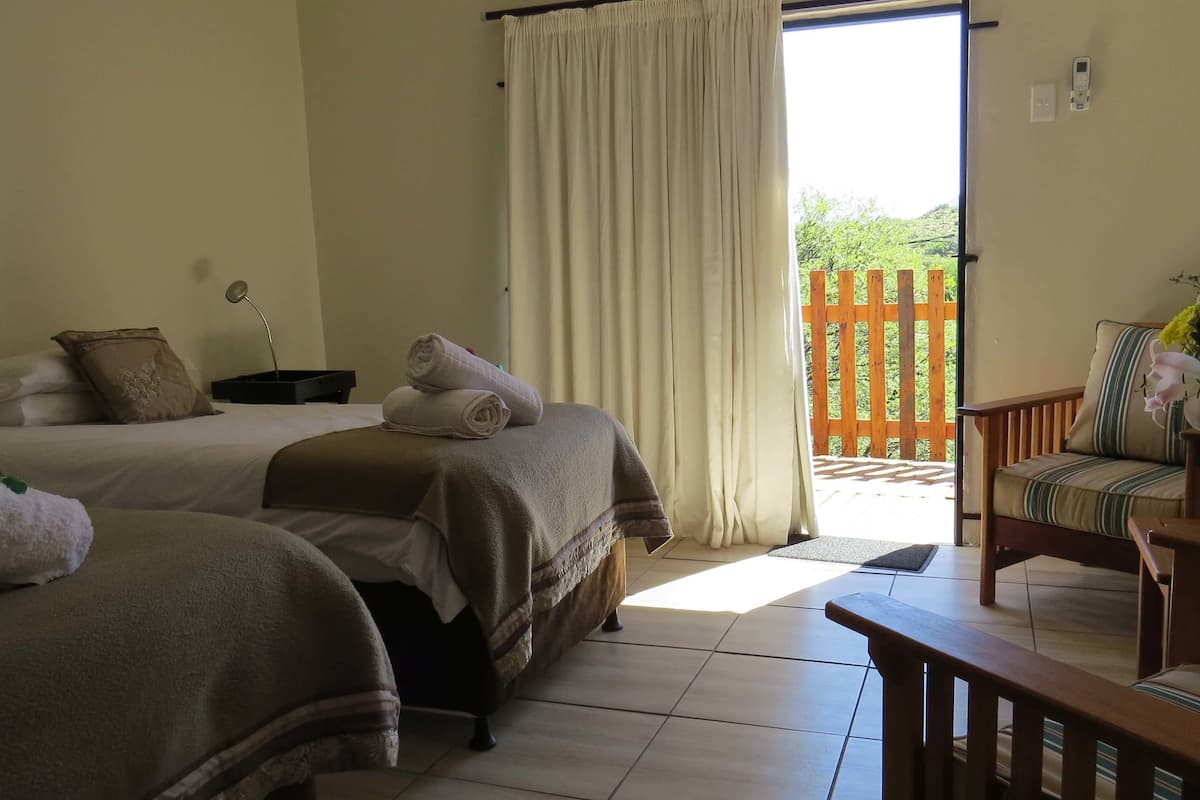 Een hotelkamer met twee bedden, een houten bankje, een stoel, een tafel, een vaas met bloemen en uitzicht op een houten terras en groen buiten.