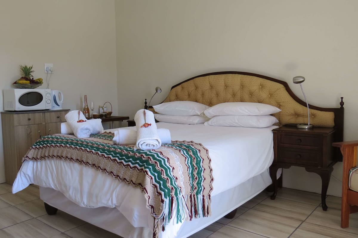 Een net opgemaakt bed met witte lakens, een decoratieve deken en opgerolde handdoeken. Er staat een magnetron op een houten kastje, een nachtkastje met een lamp, en een bordje aan de muur.