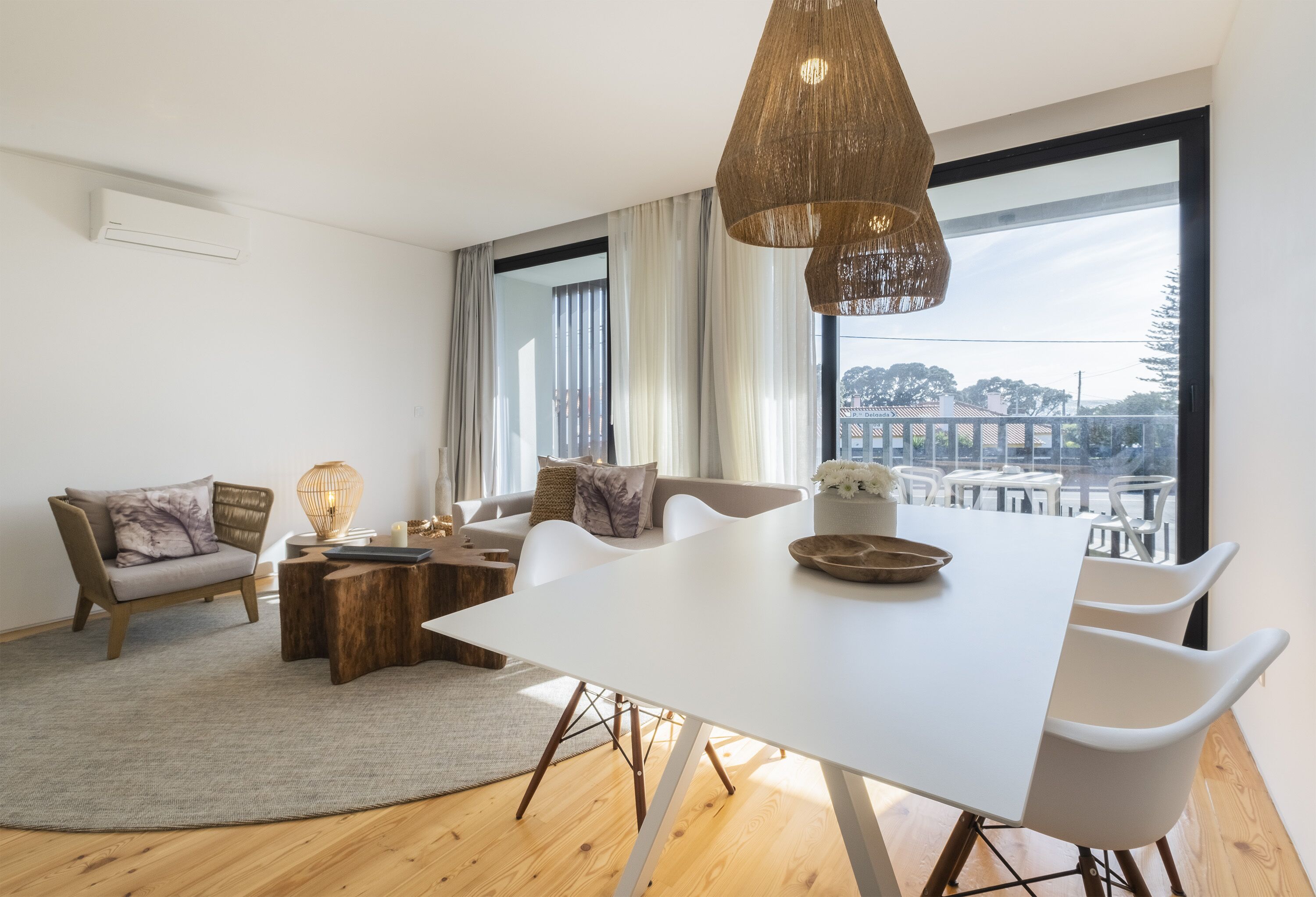 Ein modernes Wohnzimmer mit einem weißen Esstisch, weißen Stühlen, einer Couch und einem großen Fenster mit Blick auf Gebäude.