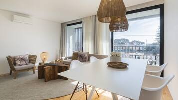 Ein modernes Wohnzimmer mit einem weißen Esstisch, weißen Stühlen, einer Couch und einem großen Fenster mit Blick auf Gebäude.