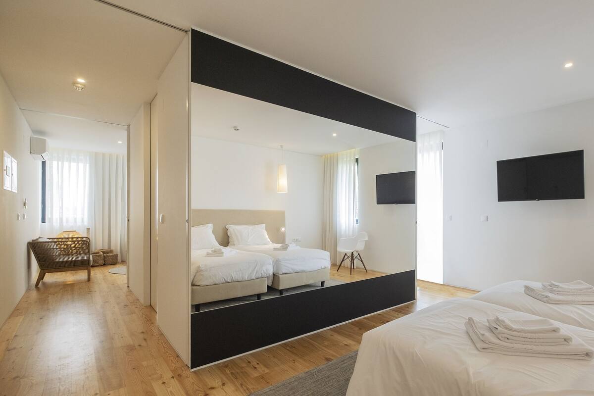 A modern hotel room with a large mirror, a bed, a TV, and a seating area with a chair and a small table.