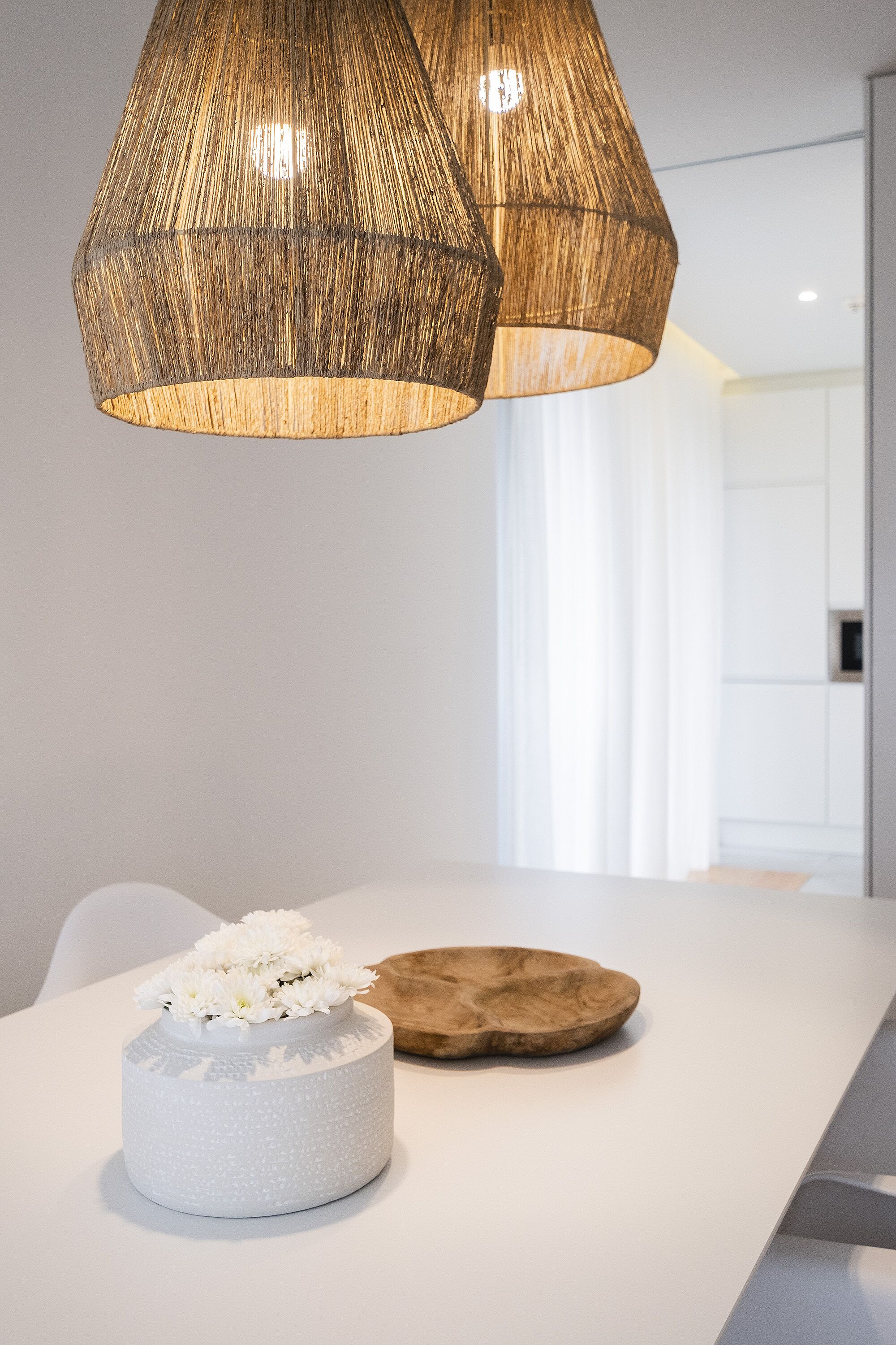 Un espace repas moderne avec une table blanche, un vase blanc à texture et orné de fleurs blanches, et un bol en bois.