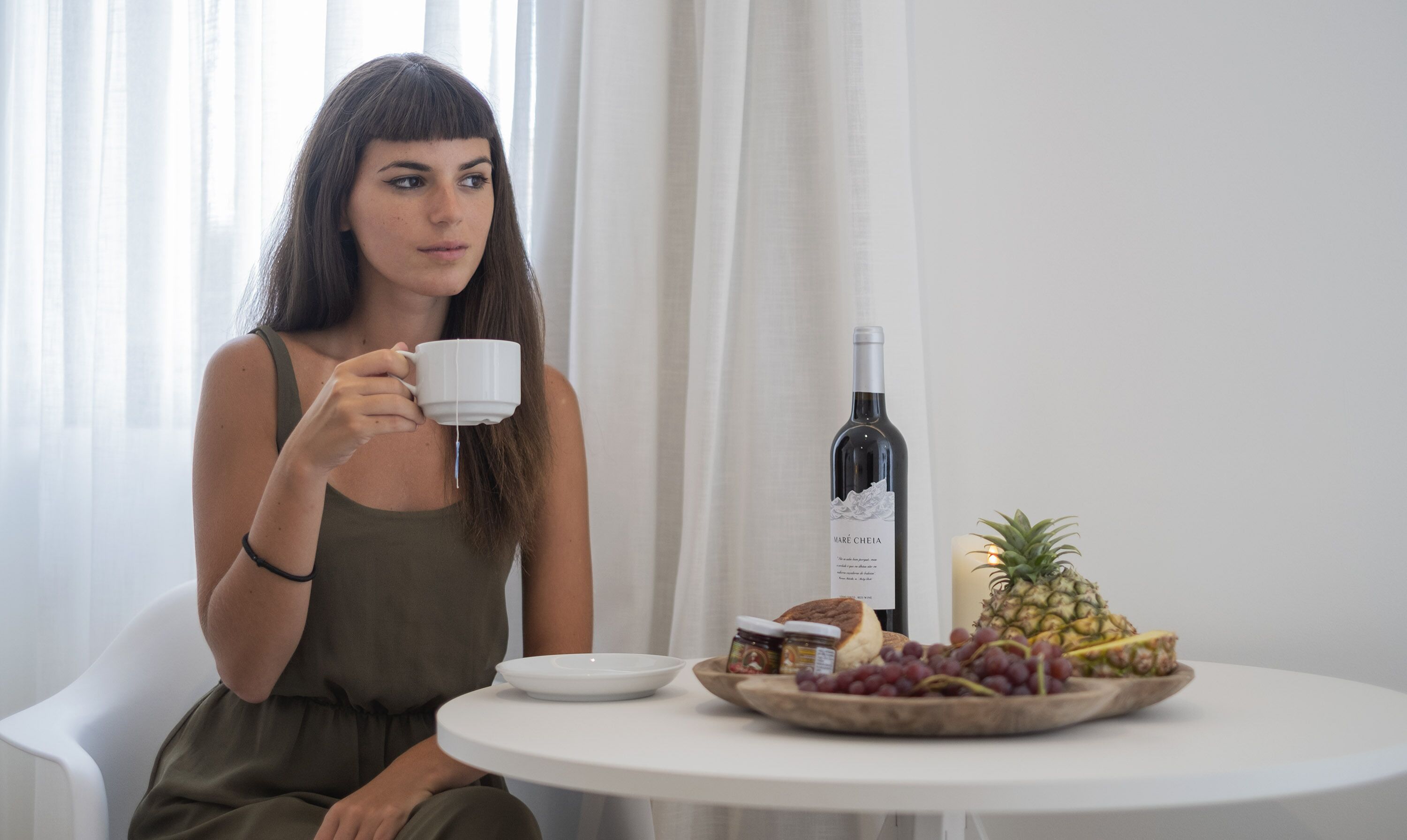 A person sitting at a round table with a white plate, a bottle of wine, a pineapple, and a bowl of grapes.