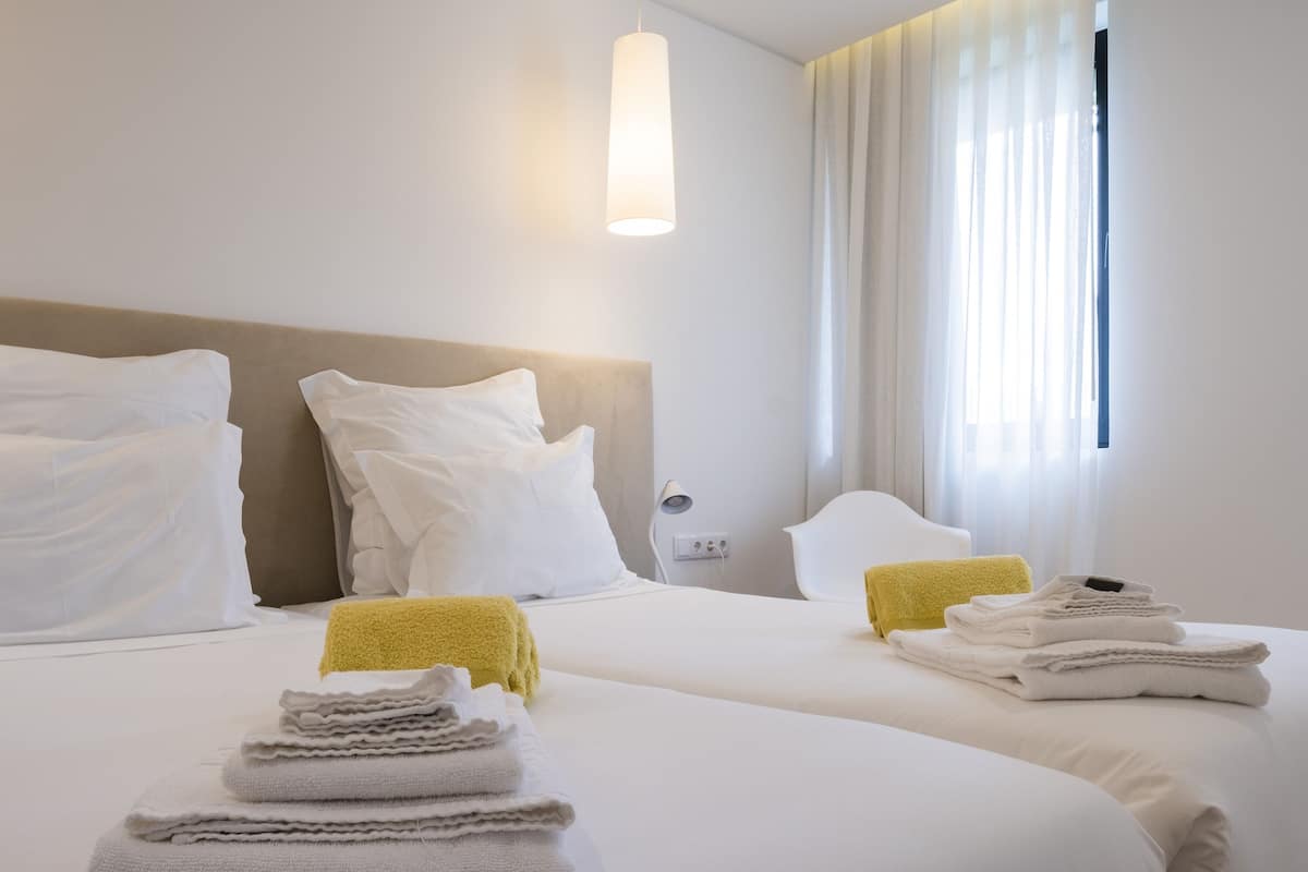 A hotel room with two beds, white linens, a headboard, a bedside lamp, and a window with curtains.