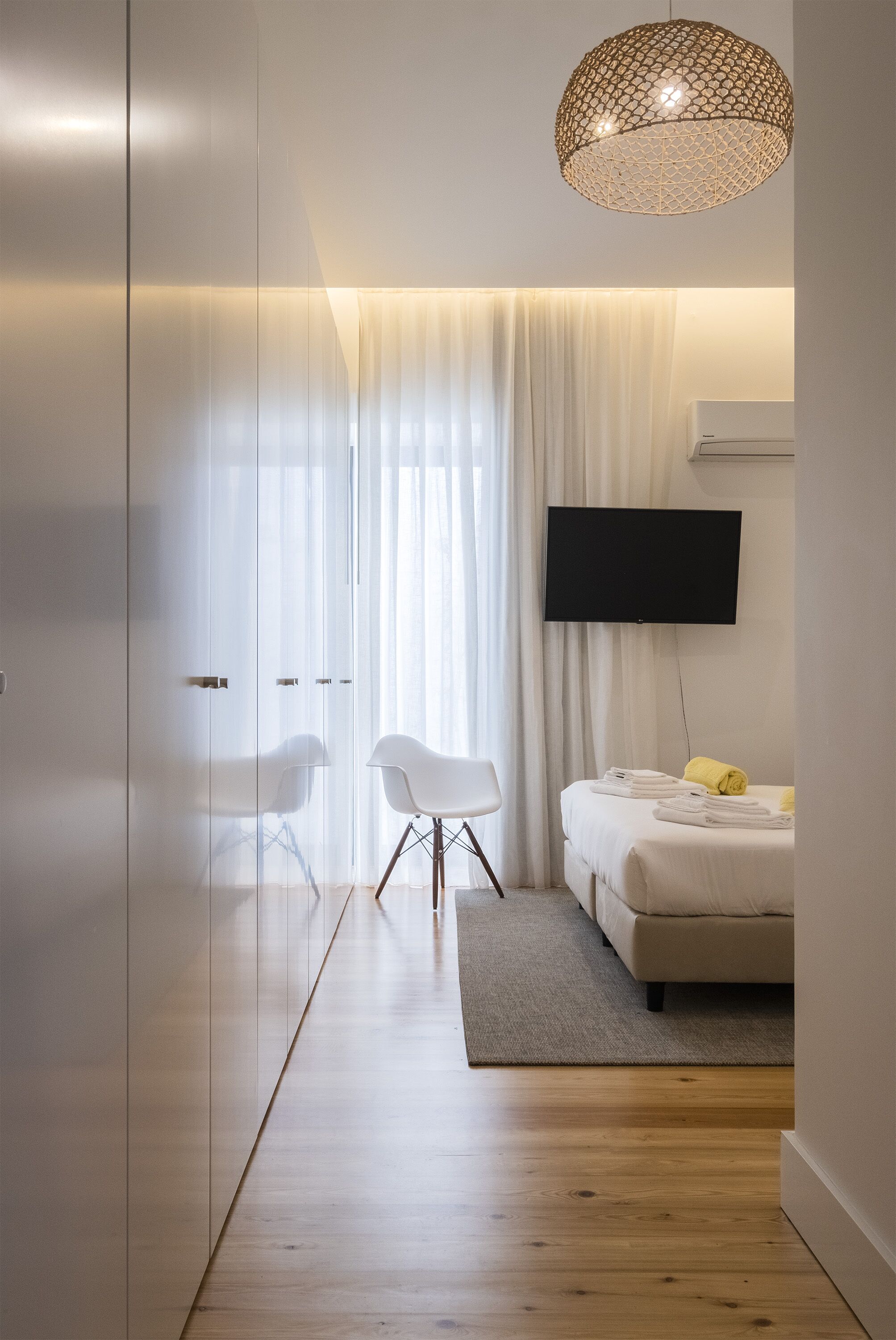 A modern hotel room with a glass partition, a flat-screen TV, a sofa, a chair, and a decorative hanging light fixture.
