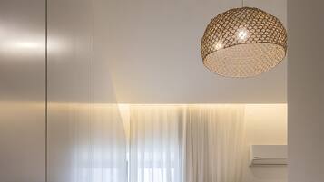 A modern hotel room with a glass partition, a flat-screen TV, a sofa, a chair, and a decorative hanging light fixture.