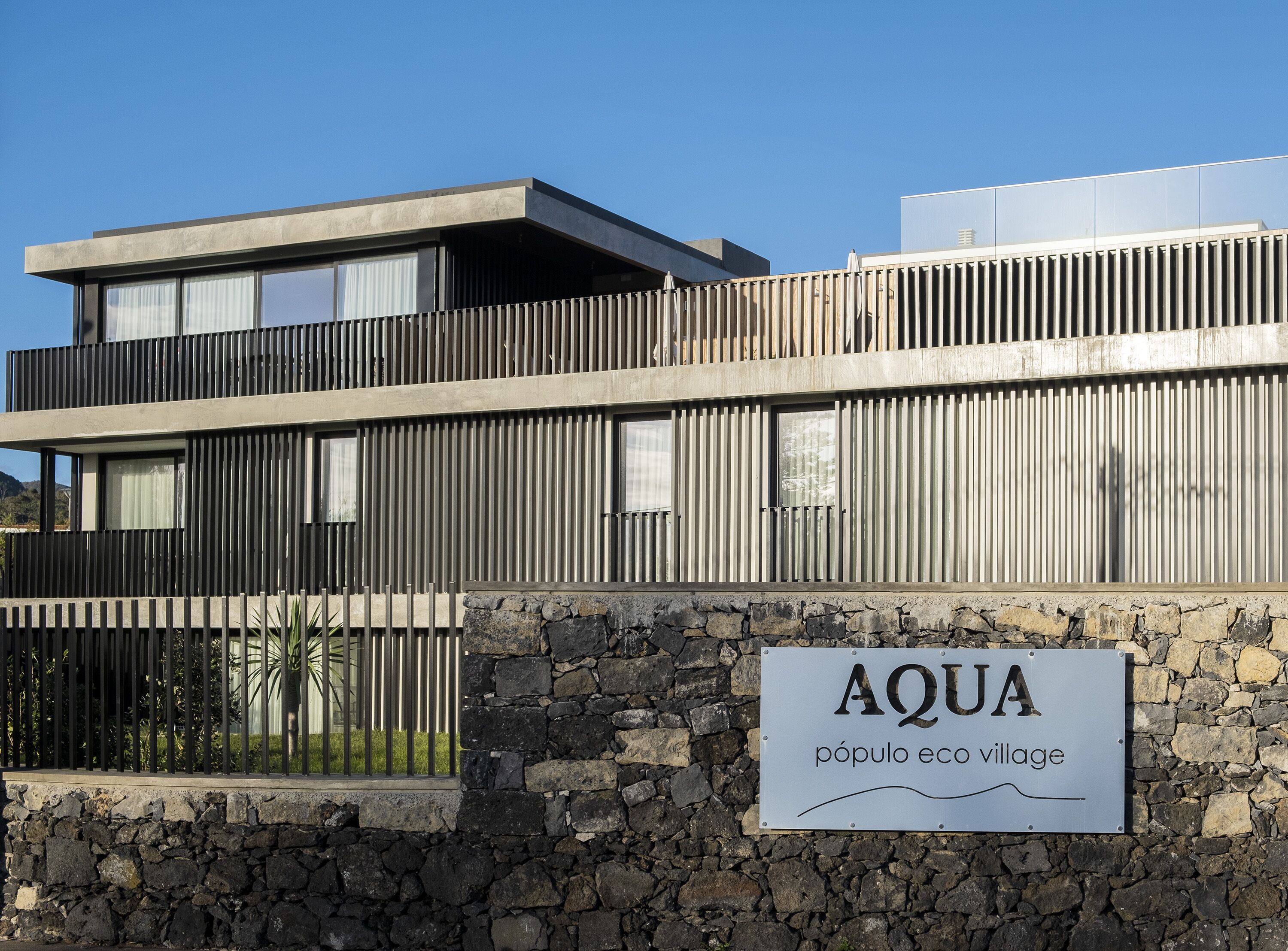 Een modern gebouw met een stenen muur en een bord waarop staat: "AQUA pueblo eco village".