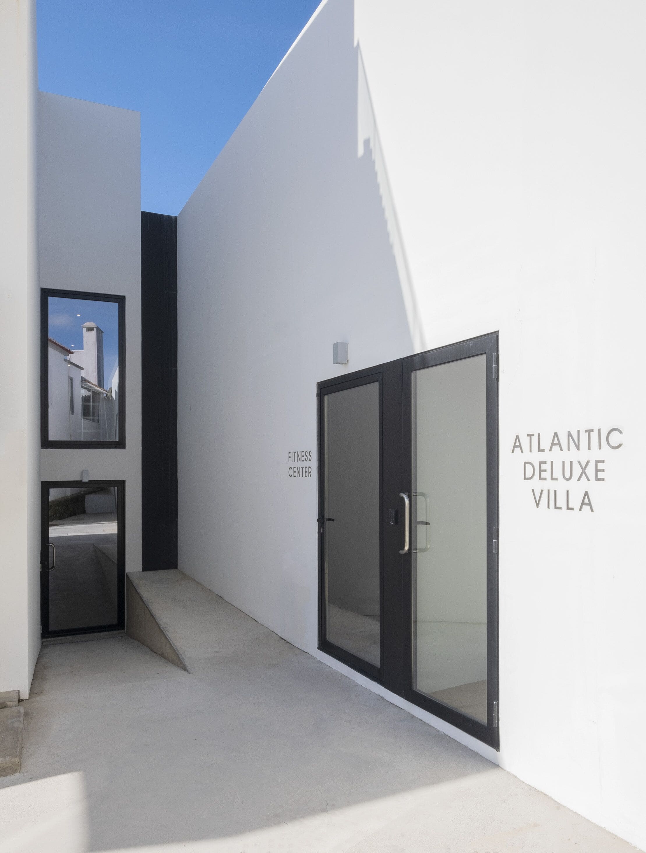 A modern building entrance with a glass door labeled "Atlantic Deluxe Villa," a small staircase, and a wall-mounted sign reading "Fitness Center."