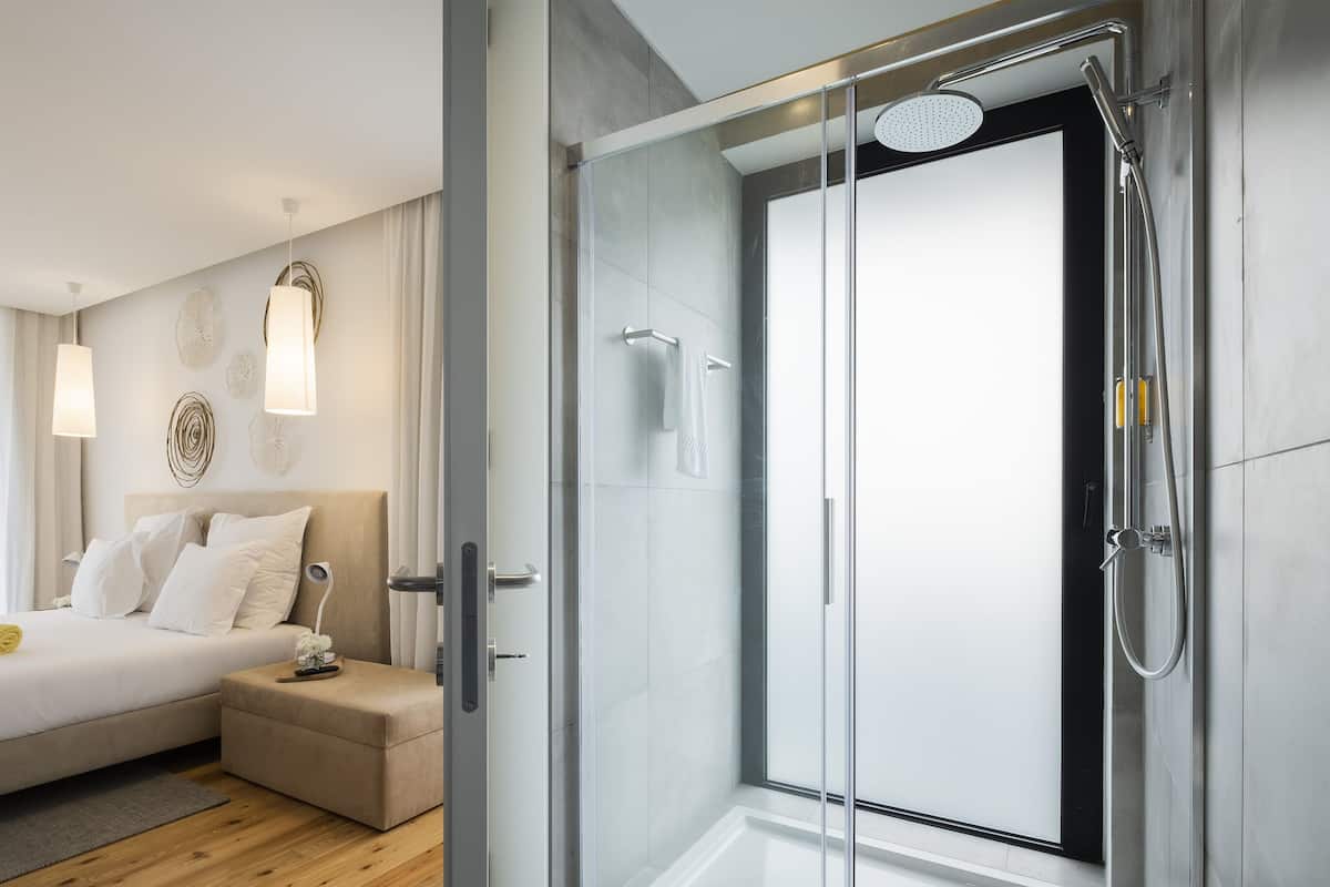 A modern hotel room with a glass-enclosed shower, a bed with white linens, and a bedside table with a vase.