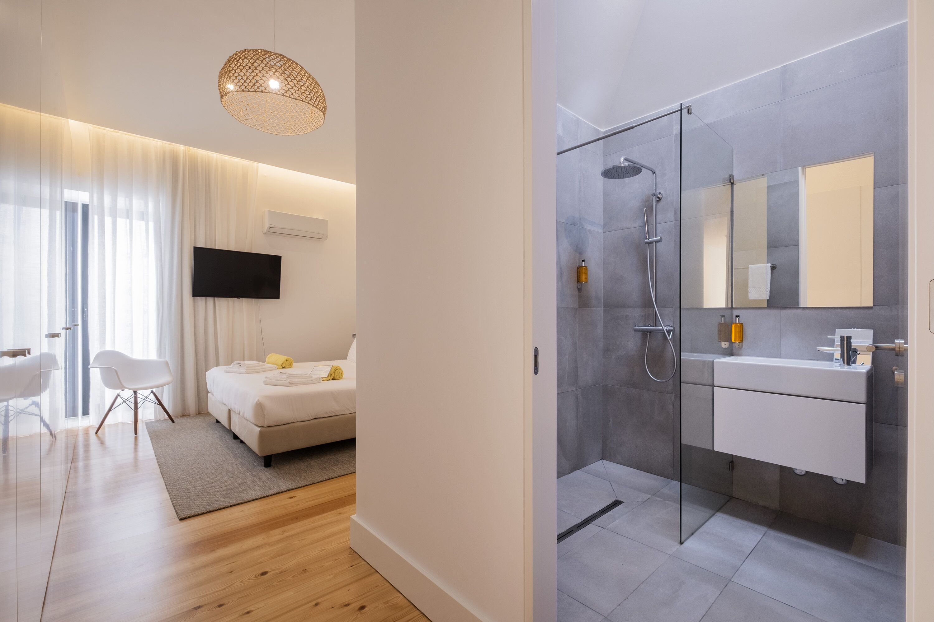 A modern hotel room with a bed, a TV, a glass shower, and a bathroom with a mirror and sink.