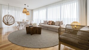 A modern living room with a round rug, a wooden coffee table, a wicker chair, a sofa with cushions, a dining table with chairs, and a hanging light fixture. - Aqua - Pópulo Eco Village (Ponta Delgada)