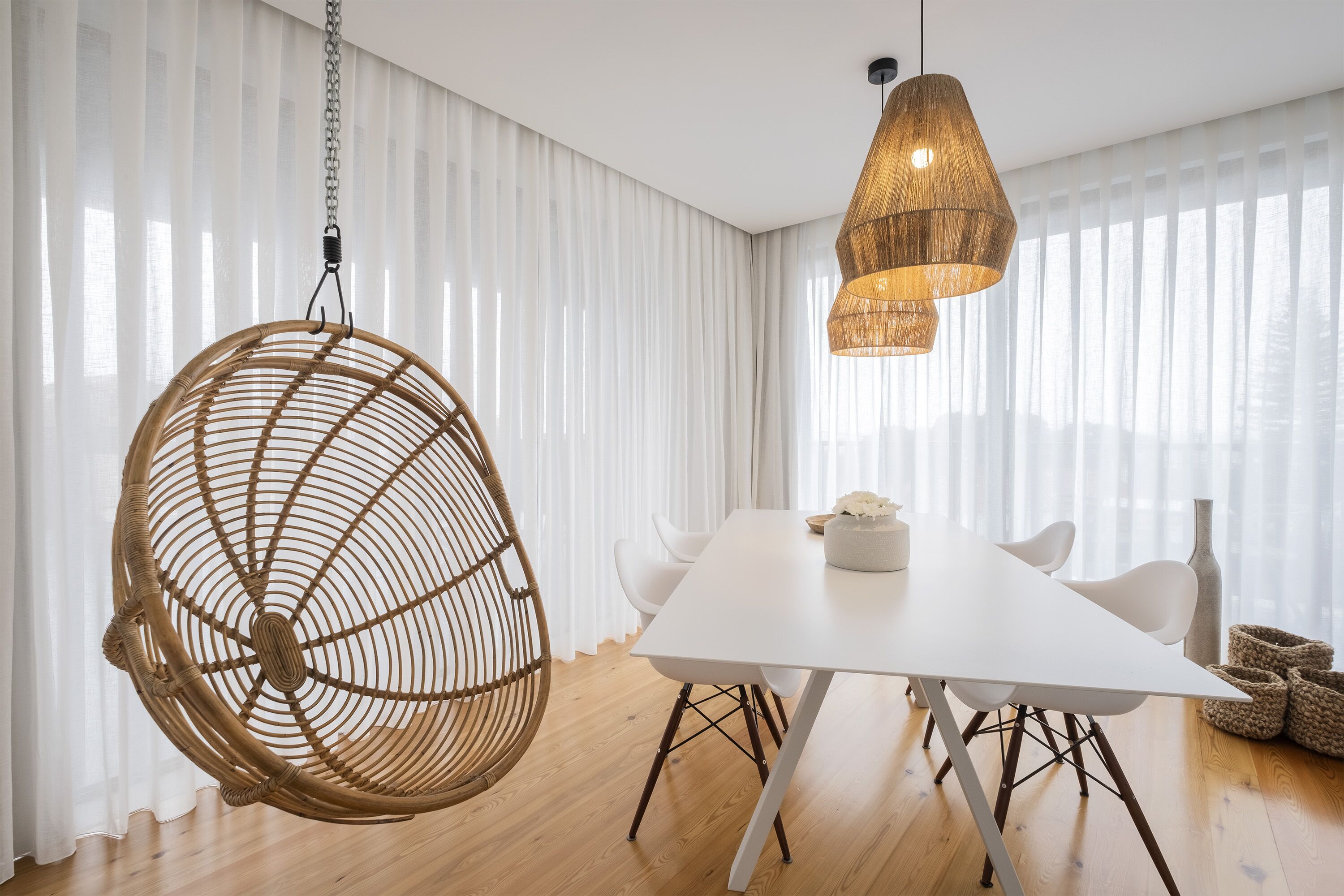 Uma área de refeições moderna com uma mesa branca, cadeiras brancas, uma cadeira de ratã suspensa e uma luminária pendente trançada.