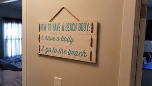 A wooden sign with a list of steps to achieve a beach body, mounted on a beige wall in a room with a bed and a window with curtains.