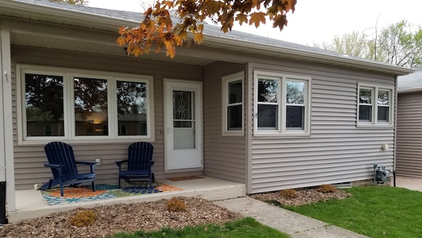 A single-story house with a front porch, two blue Adirondack chairs, and a colorful rug.