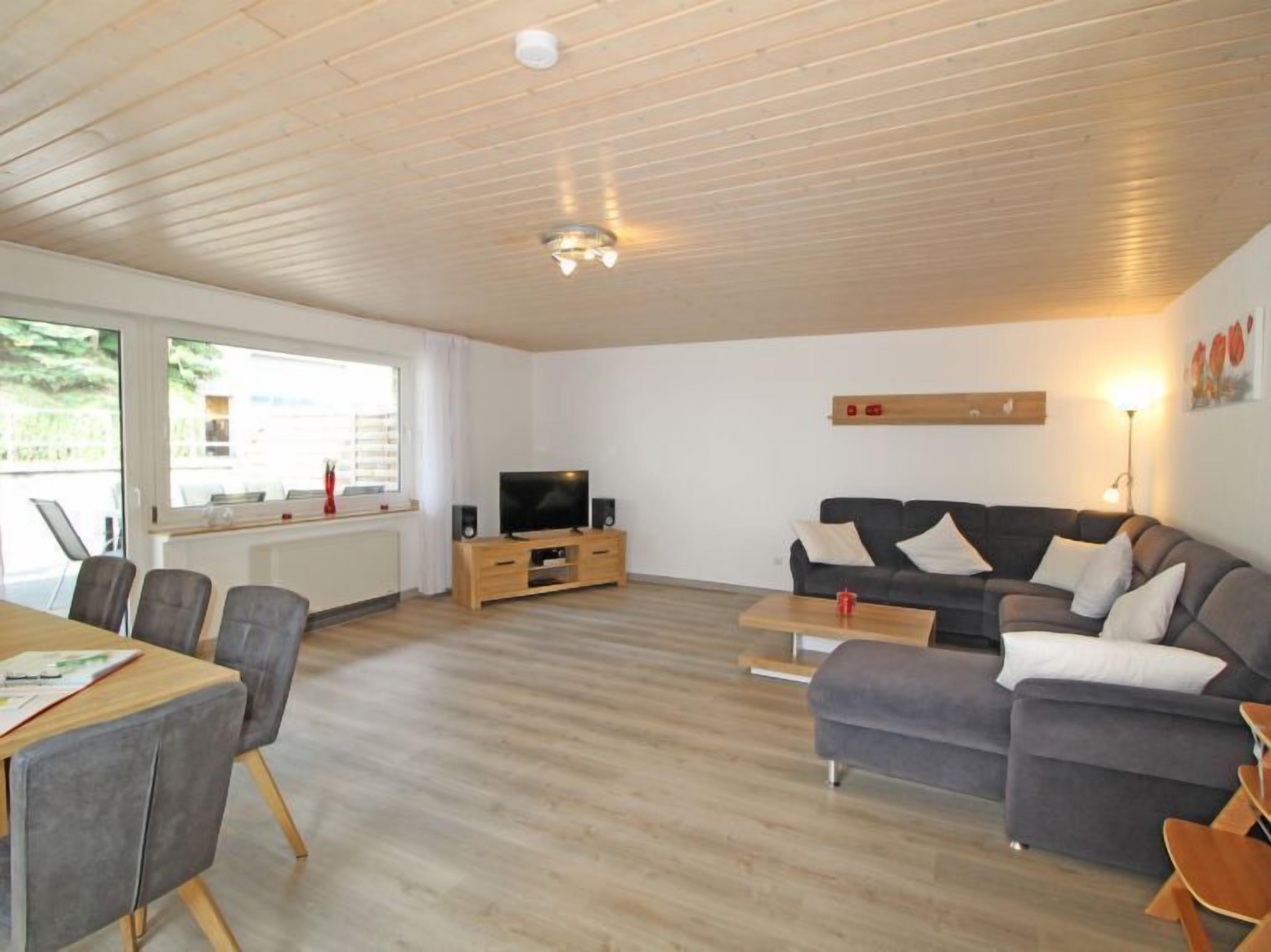 A modern living room with a wooden ceiling, a grey sofa, a wooden TV stand, and a dining area with chairs and a table.