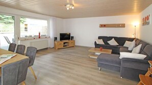 Een moderne woonkamer met een houten plafond, een grijze bank, een houten tv-meubel en een eethoek met stoelen en een tafel.