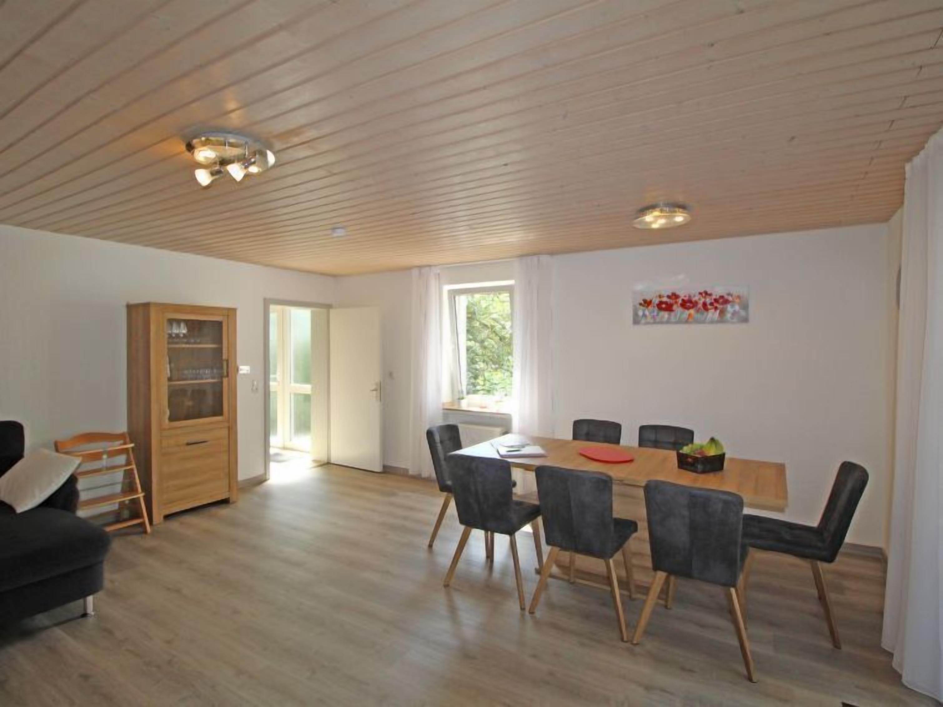 A spacious room with a wooden ceiling, a dining table with chairs, a wooden cabinet, and a window with a view of greenery.