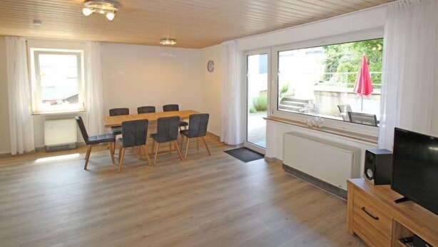 Een ruime kamer met een houten vloer, een eettafel met stoelen, een televisie op een houten dressoir en grote ramen met gordijnen.