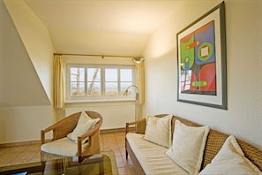 Una habitación con techo alto, una ventana con cortinas, un sofá de mimbre, una silla y un cuadro abstracto enmarcado.