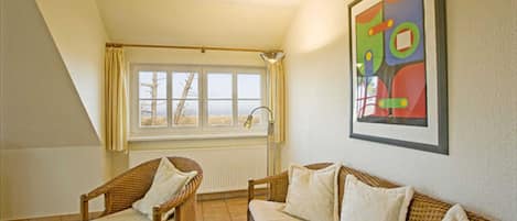 Una habitación con techo alto, una ventana con cortinas, un sofá de mimbre, una silla y un cuadro abstracto enmarcado.