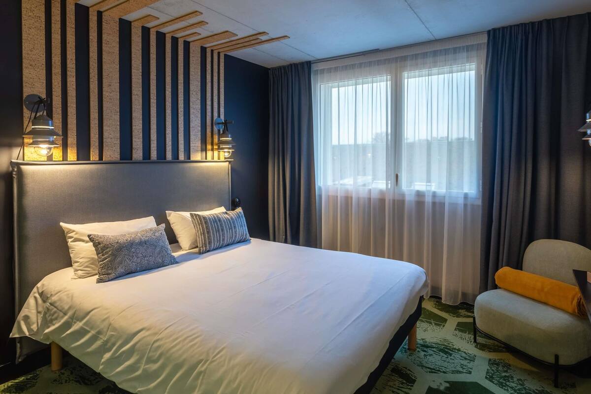 A hotel room with a large bed, a headboard with vertical slats, a window with sheer curtains, a chair with a yellow cushion, and a patterned rug.