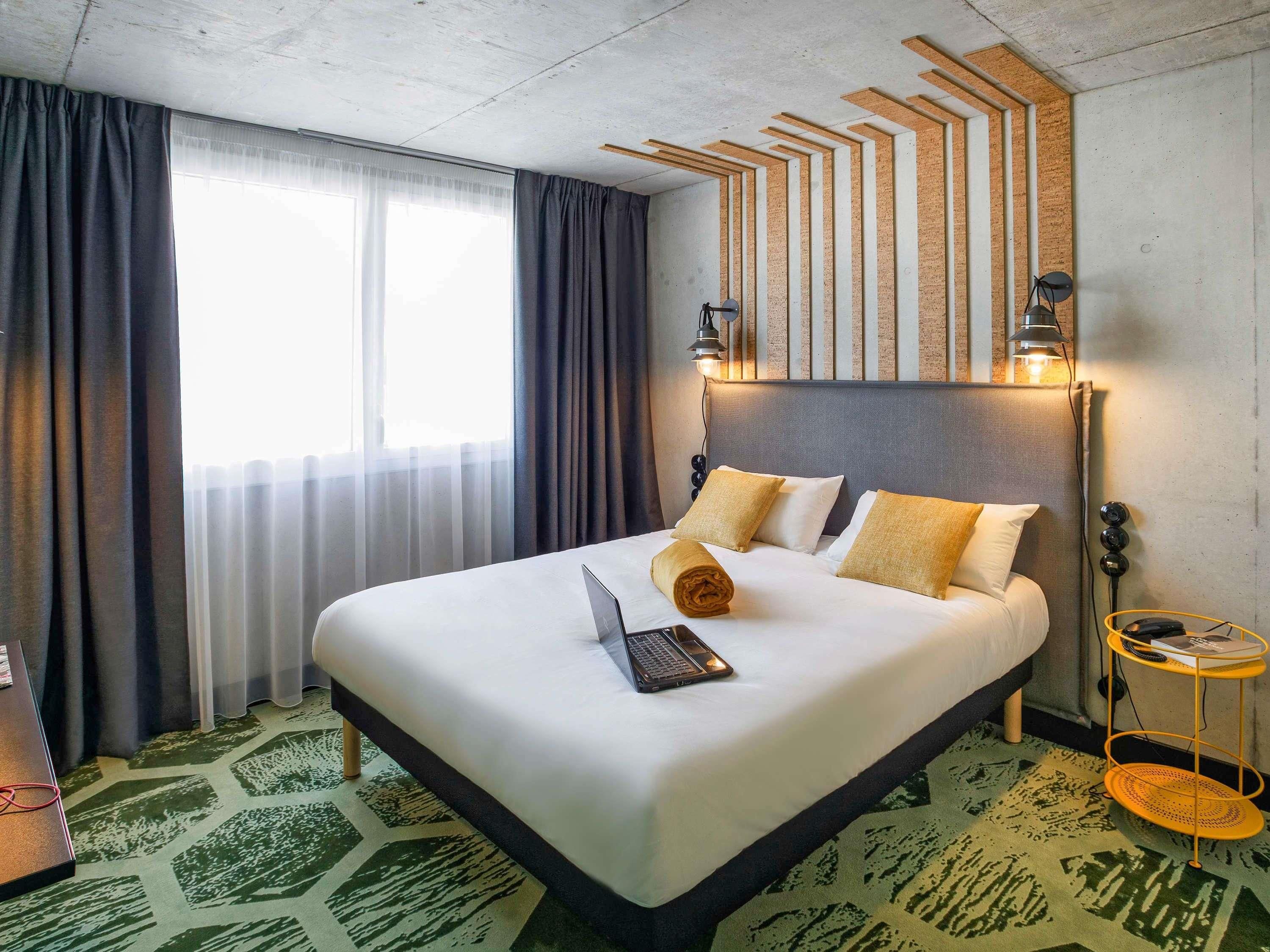 A hotel room with a bed, a laptop, a bedside table with a lamp, and a view of the outdoors.