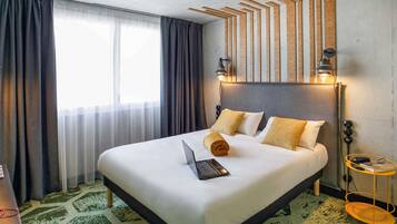 A hotel room with a bed, a laptop, a bedside table with a lamp, and a view of the outdoors.