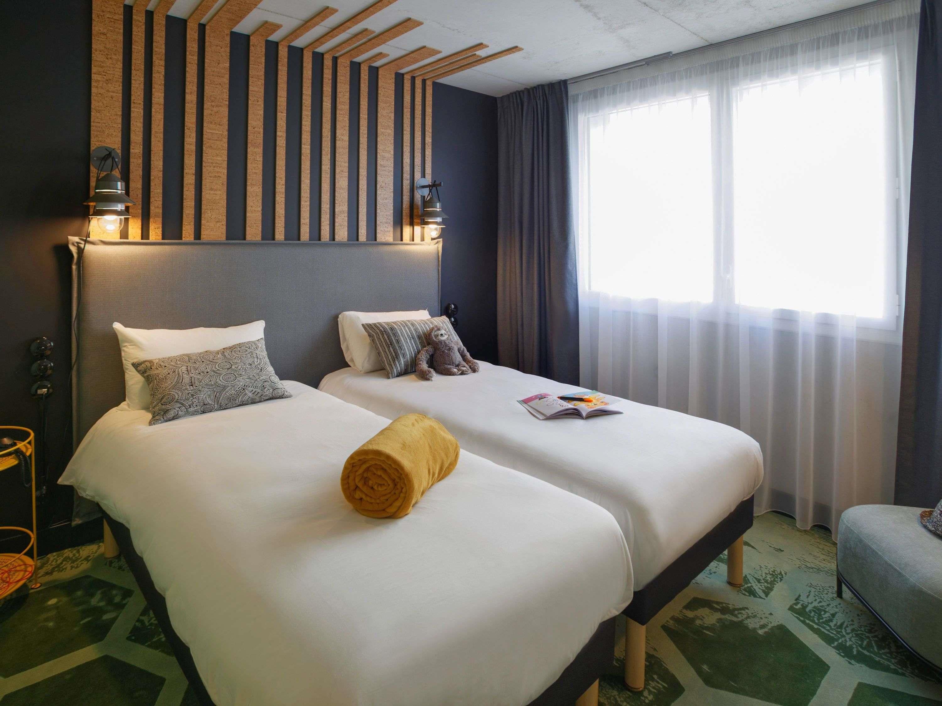 A hotel room with two beds, a wooden slat headboard, a large window with curtains, and a green patterned carpet.