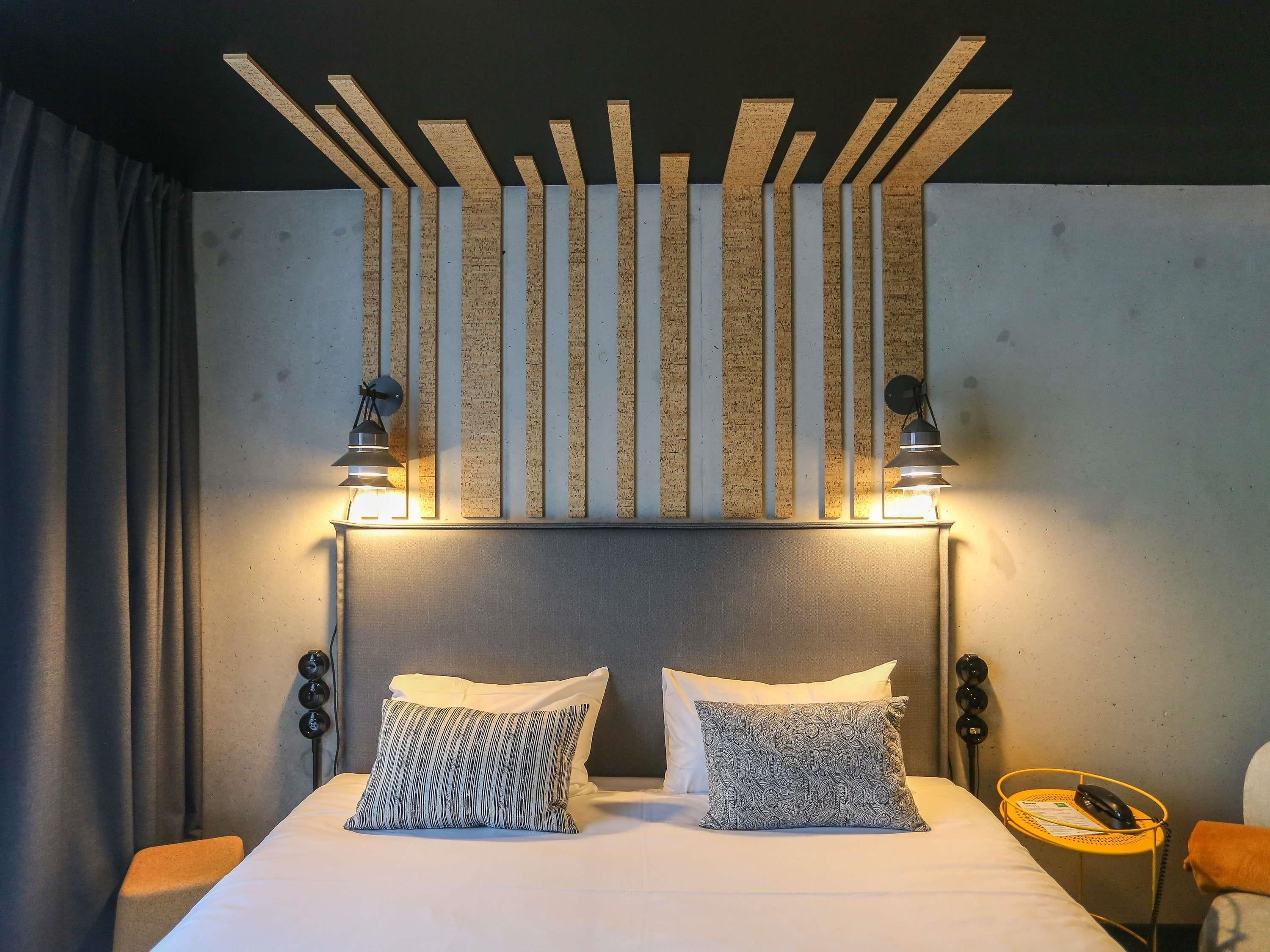 A hotel room with a bed, two pillows, a headboard, a nightstand, a lamp, and a wall-mounted light fixture.