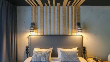 A hotel room with a bed, two pillows, a headboard, a nightstand, a lamp, and a wall-mounted light fixture.