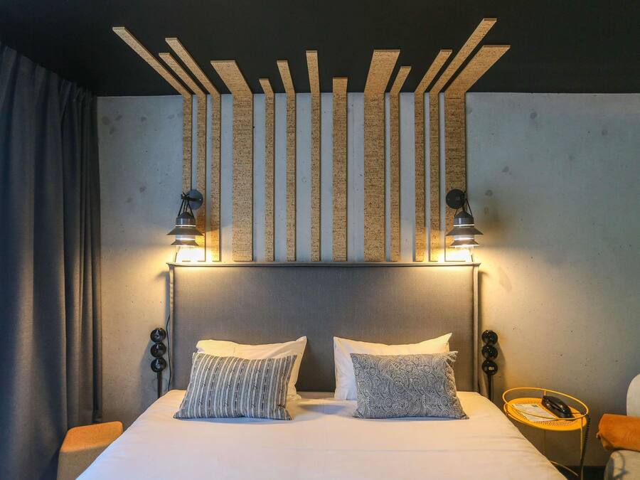 A hotel room with a bed, two pillows, a headboard, a nightstand, a lamp, and a wall-mounted light fixture.