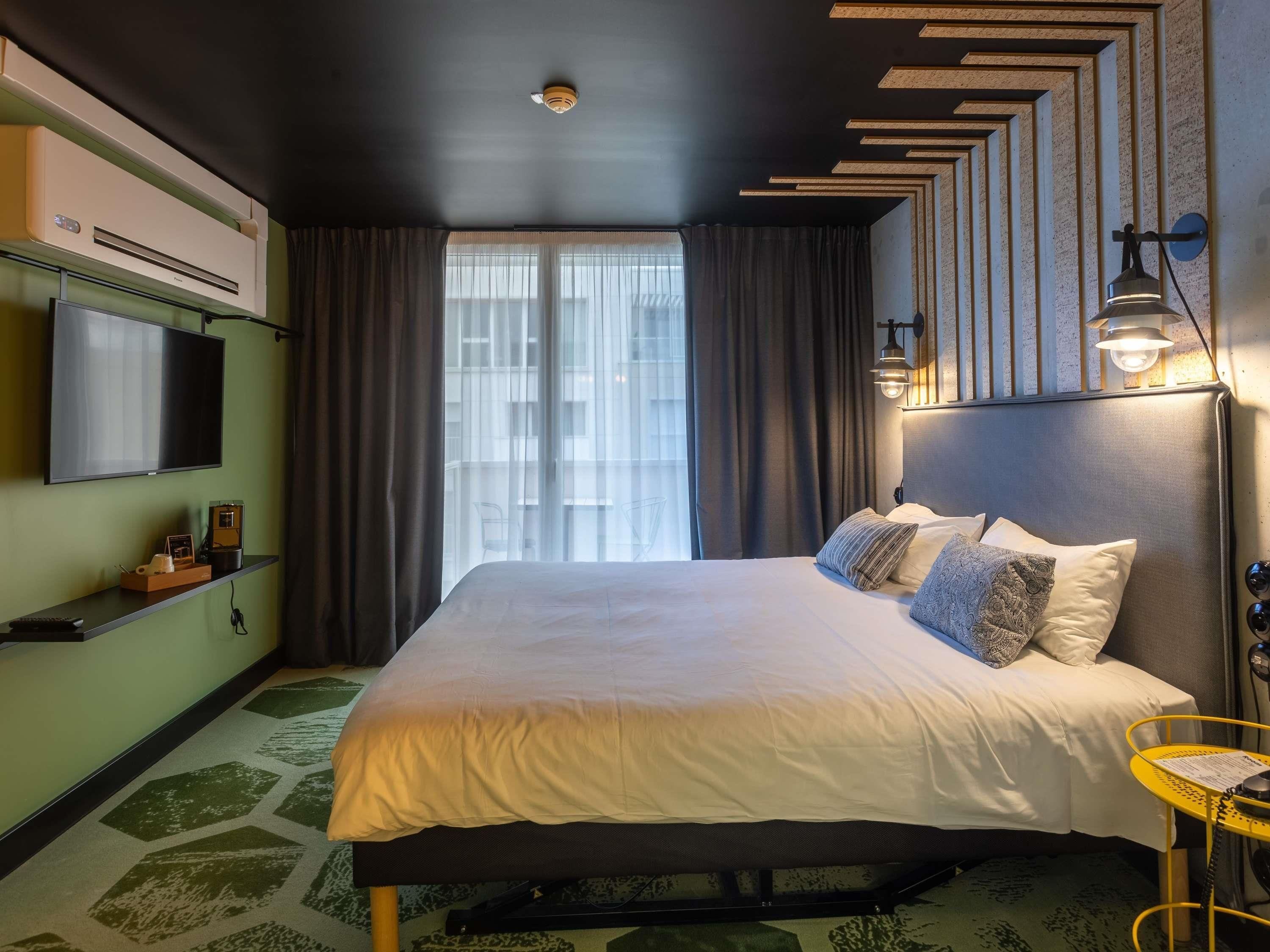 A modern hotel room with a large bed, a flat-screen TV, a coffee machine, and a view of the city through the window.